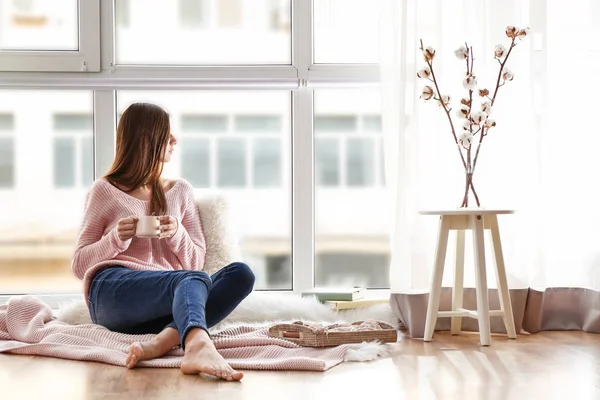 Ung kvinna dricka varmt kaffe nära fönster — Stockfoto