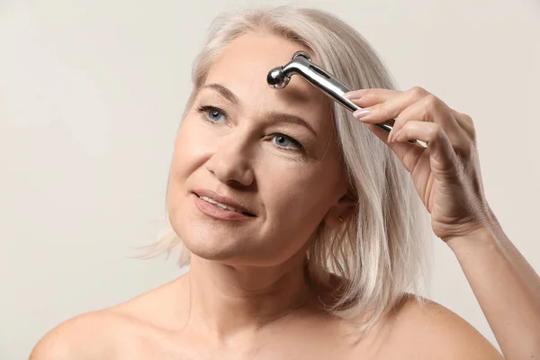 Mulher madura com ferramenta de massagem facial no fundo claro — Fotografia de Stock