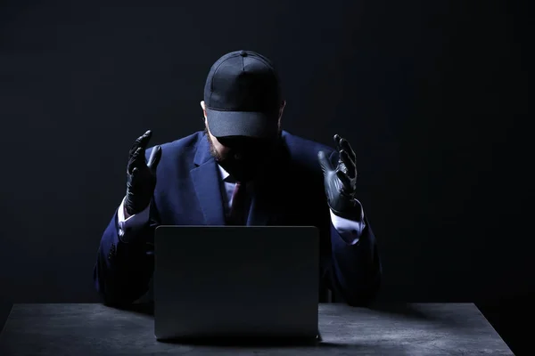 Professionele hacker met laptop zittend aan tafel op donkere achtergrond — Stockfoto