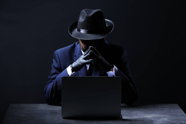 Professional hacker with laptop sitting at table on dark background