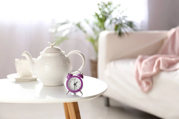 Relógio despertador, bule e xícara na mesa na sala de luz — Fotografia de Stock