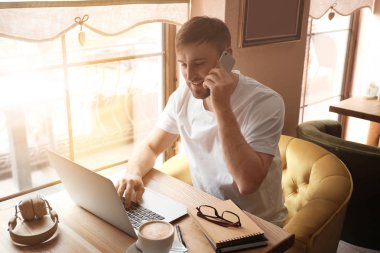 Kafede dizüstü bilgisayarda çalışırken cep telefonuyla konuşan genç serbest meslek