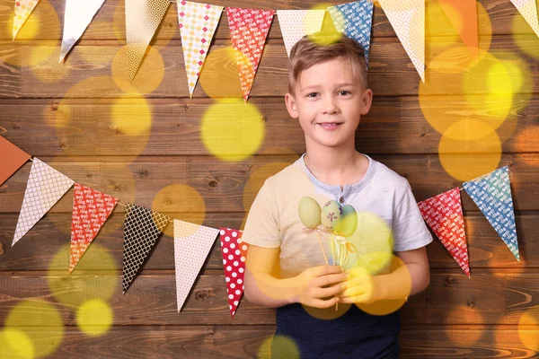 Lindo niño pequeño con huevos de Pascua cerca de la pared de madera decorada con banderines de fiesta —  Fotos de Stock