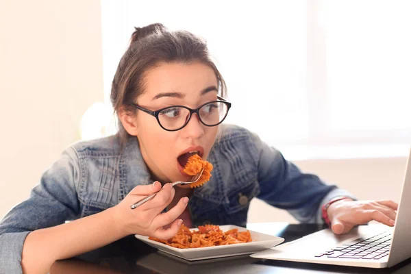 Молода жінка їсть смачну пасту, використовуючи ноутбук у приміщенні — стокове фото