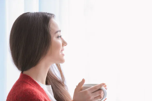 家窗边的漂亮年轻女子, 旁边有杯热茶。 — 图库照片