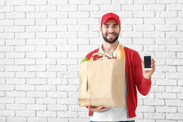 Доставщик держит бумажный пакет с едой и телефоном на фоне кирпичной стены — стоковое фото