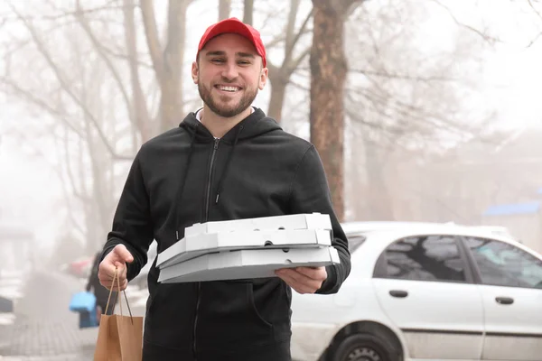 Young man with cardboard pizza boxes and paper bag outdoors. Food delivery service — Stock Photo, Image