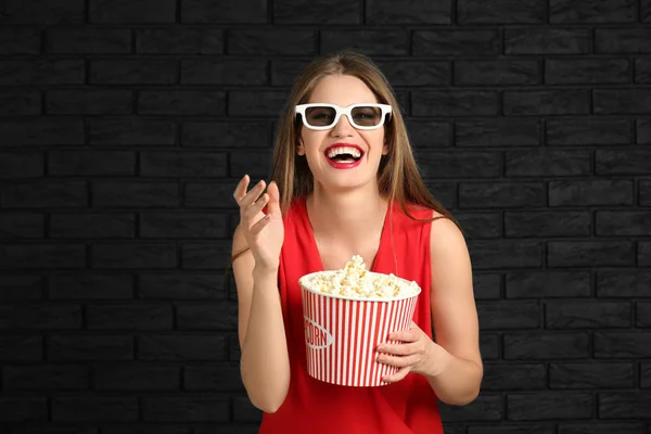 Schöne junge Frau mit einer Tasse Popcorn und 3D-Kinobläsern gegen eine dunkle Backsteinwand — Stockfoto