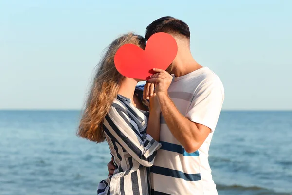 Linda pareja joven sosteniendo el corazón rojo en la orilla del mar —  Fotos de Stock