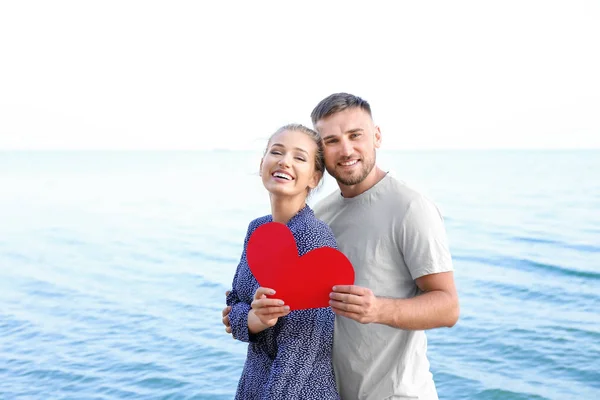 Cute young couple with red heart on sea shore — Stock Photo, Image