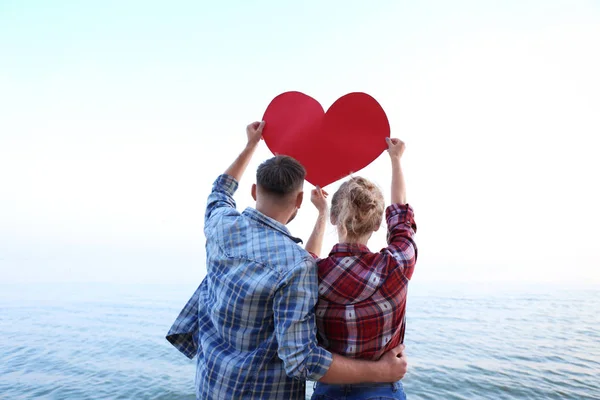 Linda pareja joven con corazón rojo en la orilla del mar —  Fotos de Stock