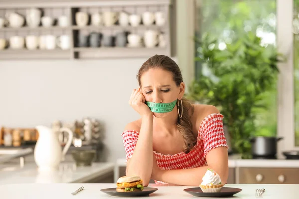 Emotional woman with measuring tape around her mouth and unhealthy food in kitchen. Diet concept — Stock Photo, Image