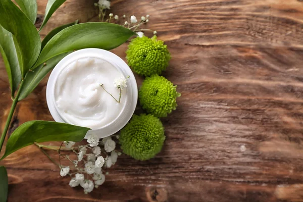 Jar with body cream and flowers on wooden table, top view