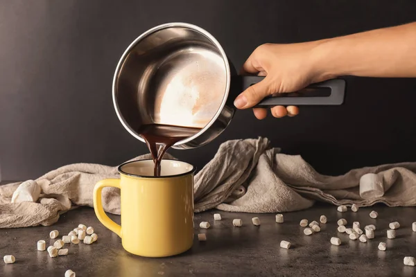 Woman pouring hot chocolate from pot into metal cup on dark table — Stock Photo, Image