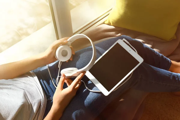 Mladá žena s tabletovým počítačem a sluchátka sedí u okna doma — Stock fotografie