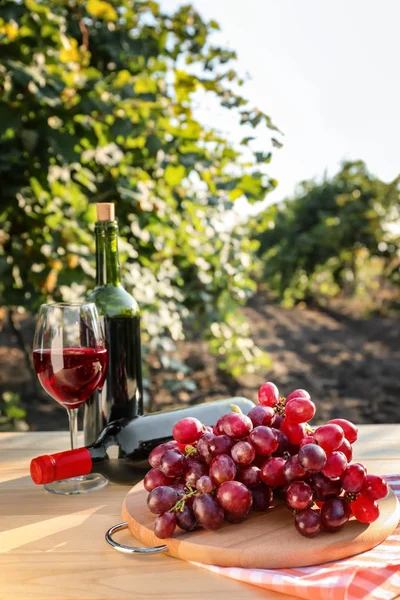 Свежий виноград с бутылками и бокалом красного вина на деревянном столе в винограднике — стоковое фото