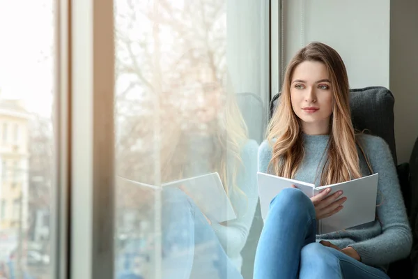 Schöne junge Frau liest Buch, während sie auf der Fensterbank sitzt — Stockfoto