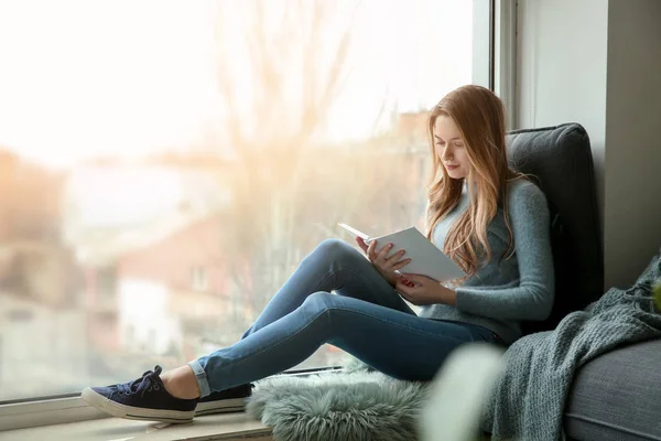 Hermosa joven leyendo libro mientras está sentado en el alféizar de la ventana — Foto de Stock