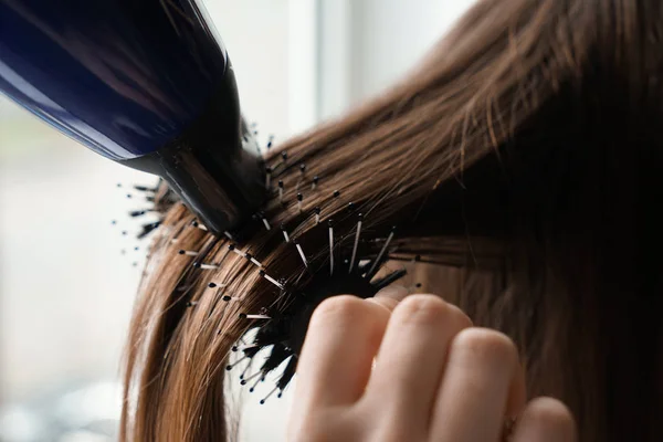 Cabeleireiro com secador de cabelo escovando cabelos longos de mulher jovem no salão, close-up — Fotografia de Stock