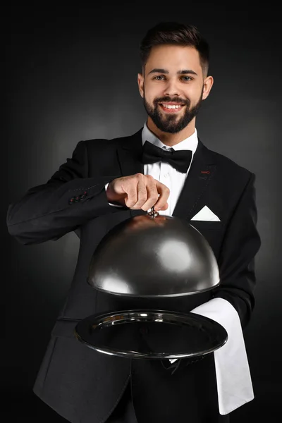 Handsome waiter with tray and cloche on dark background — Stock Photo, Image