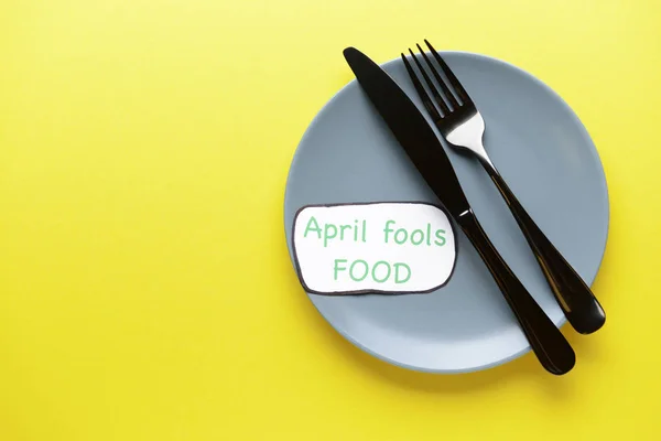 Sheet of paper with words "April fools food", plate and cutlery on color background — Stock Photo, Image