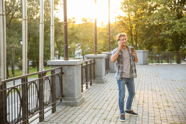 Handsome young man with coffee talking on mobile phone in park — Stock Photo, Image