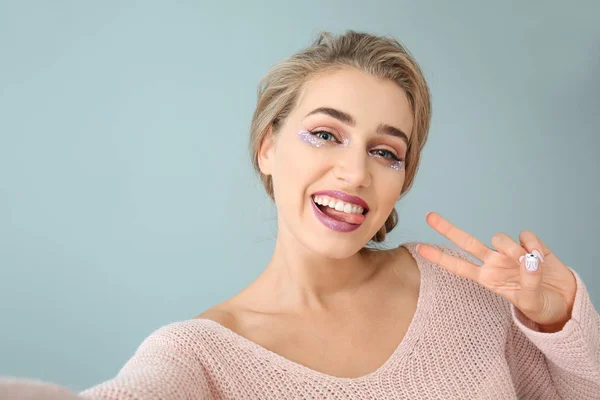 Beautiful young woman showing Victory gesture while taking selfie on color background — Stock Photo, Image