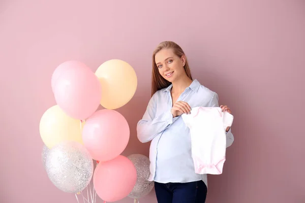 Femeie gravidă frumoasă cu haine pentru copii și baloane de aer pe fundal de culoare — Fotografie, imagine de stoc