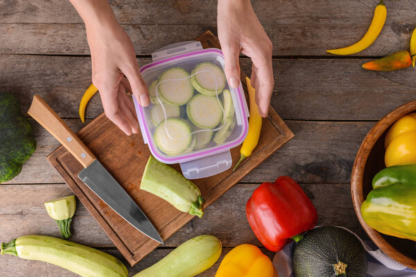 Woman holding plastic container with fresh squash for freezing on wooden table