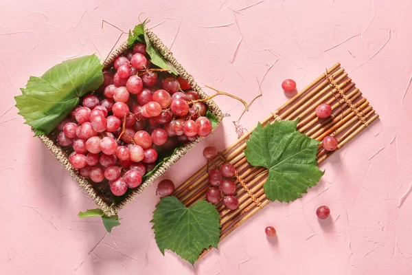 Wicker box with ripe grapes and bamboo mat on color background