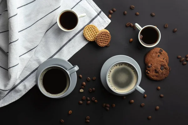 Viele Tassen mit leckerem aromatischen Kaffee und Keksen auf schwarzem Hintergrund, flache Lage — Stockfoto