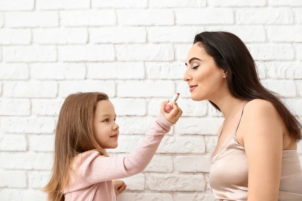 Cute little daughter applying lipstick onto mother's lips against white brick background — Stock Photo, Image