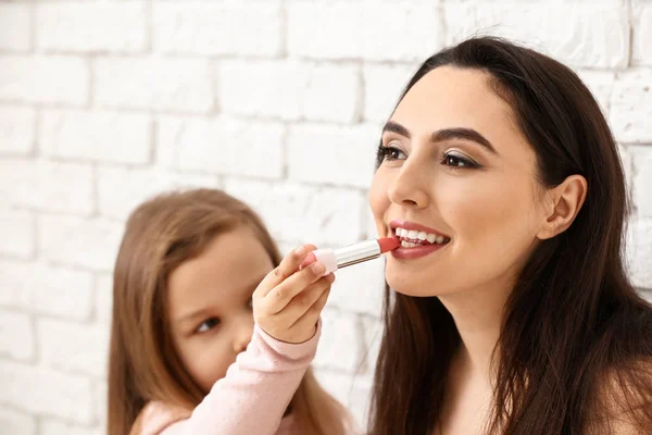 Niedliche kleine Tochter trägt Lippenstift auf die Lippen der Mutter vor weißem Backsteinhintergrund auf — Stockfoto