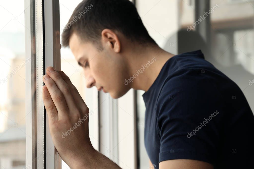 Depressed young man standing near window and thinking about suicide