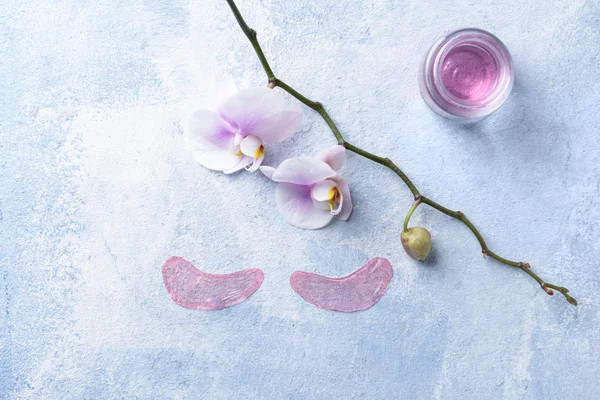 Under-eye patches with cosmetic product and orchid flowers on color background