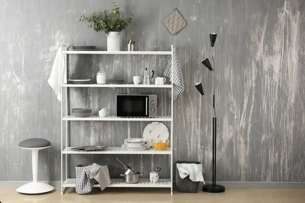 Set di piatti puliti con utensili e forno a microonde sugli scaffali vicino alla parete grigia — Foto Stock