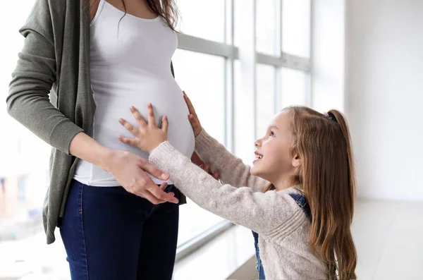 Niña con madre embarazada cerca de la ventana en casa — Foto de Stock