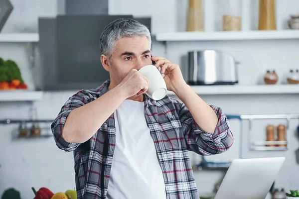 Mature man talking by phone while drinking coffee in kitchen — Stock Photo, Image