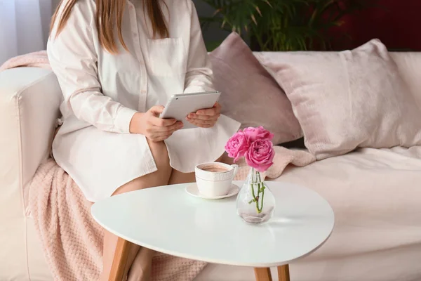 Young woman using tablet PC near table with cup of coffee at home — Stock Photo, Image