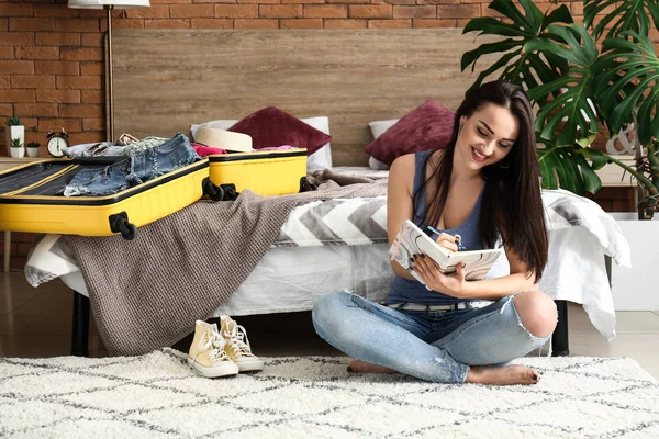 Young woman checking list of things while packing suitcase at home