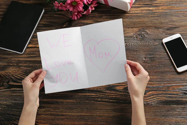 Mani femminili con biglietto di auguri per la festa della mamma su sfondo di legno — Foto Stock