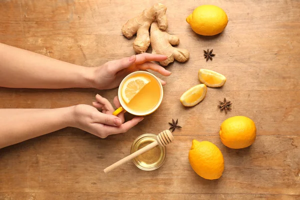 Manos femeninas con taza de sabroso té, limón y jengibre sobre fondo de madera —  Fotos de Stock