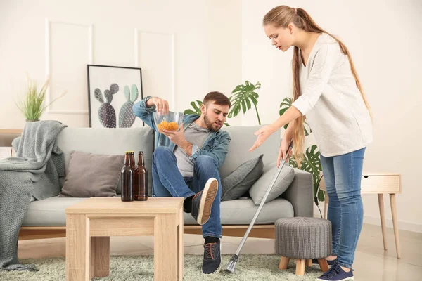 Dona de casa com escova repreendendo seu marido preguiçoso descansando no sofá — Fotografia de Stock