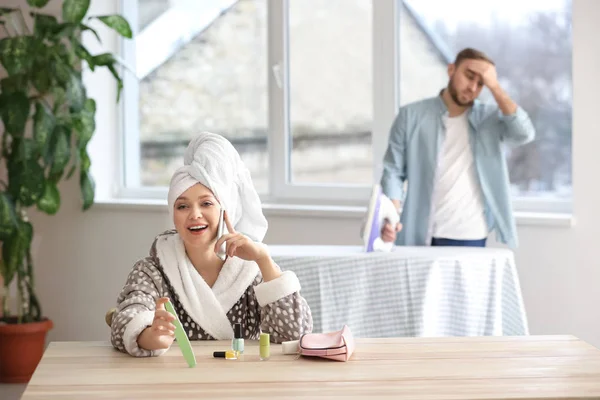 Carefree wife sitting at table while her indignant husband doing chores at home — Stock Photo, Image