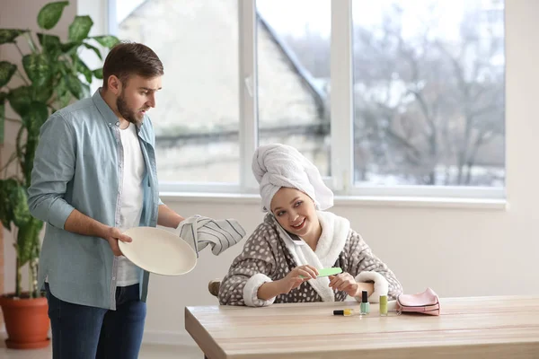 Unbekümmerte Ehefrau sitzt am Tisch, während ihr empörter Ehemann zu Hause Hausarbeiten erledigt — Stockfoto