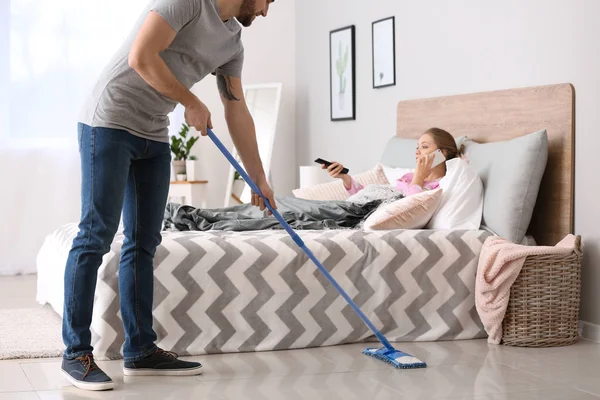 Househusband cleaning floor near lazy wife lying in bed