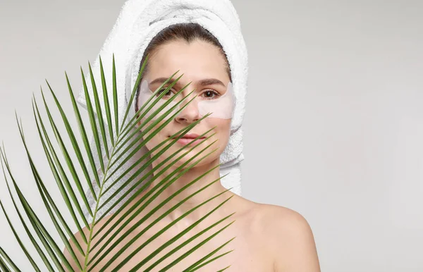 Woman with under-eye patches and tropical leaf on light background