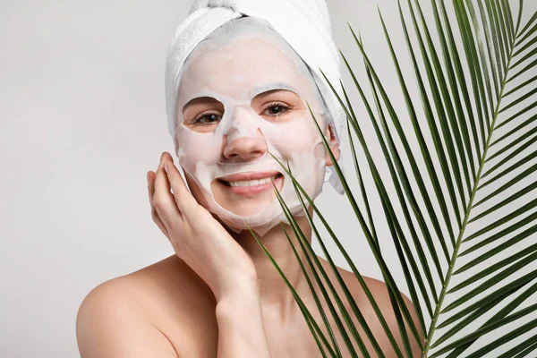 Vrouw met vel gezichtsmasker en tropisch blad op lichte achtergrond — Stockfoto
