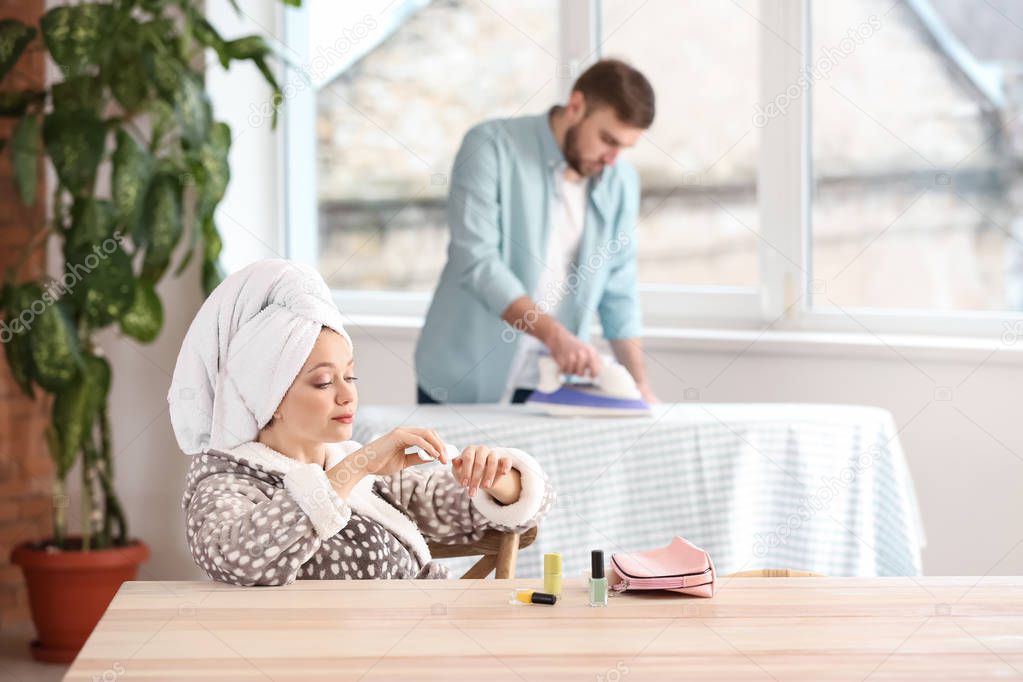 Carefree wife sitting at table while her husband doing chores at home