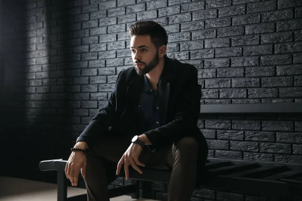 Elegantní mladý muž sedí na lavičce proti tmavě cihlová zeď — Stock fotografie